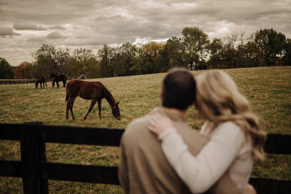 Couple cuddles beside a pasture while horse gently grazes.