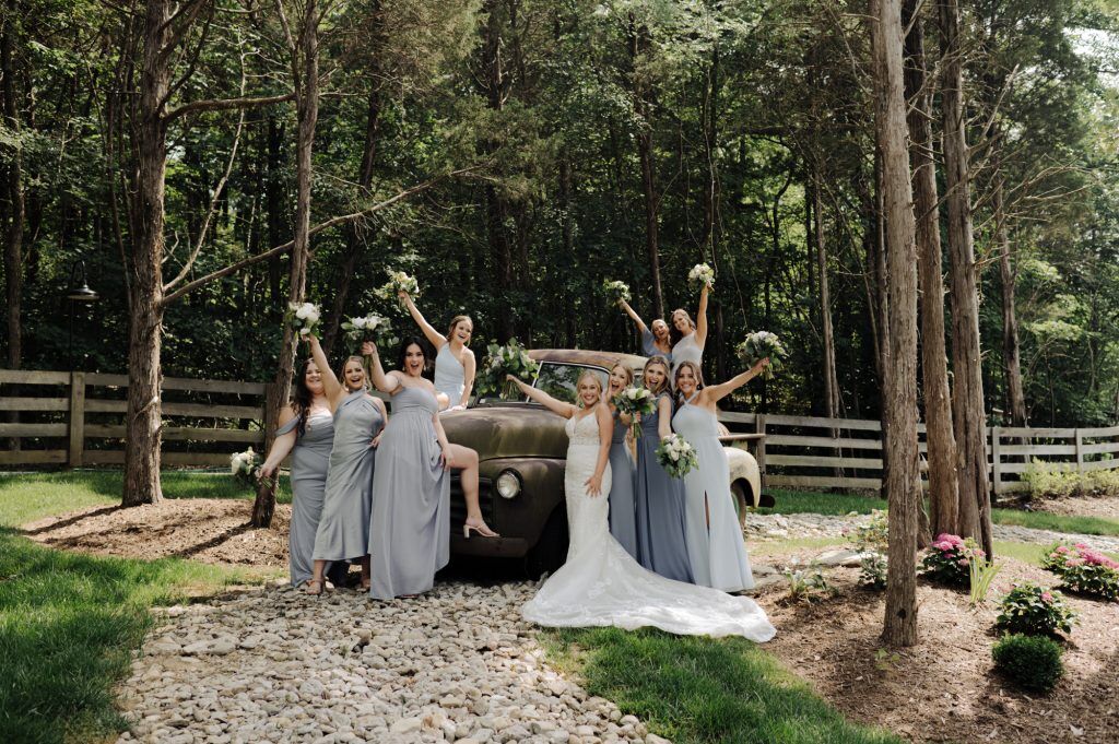 Bridesmaids and bride celebrate the recent marriage of their friend near an old car in the woods at Woods at Cedar Springs Venue